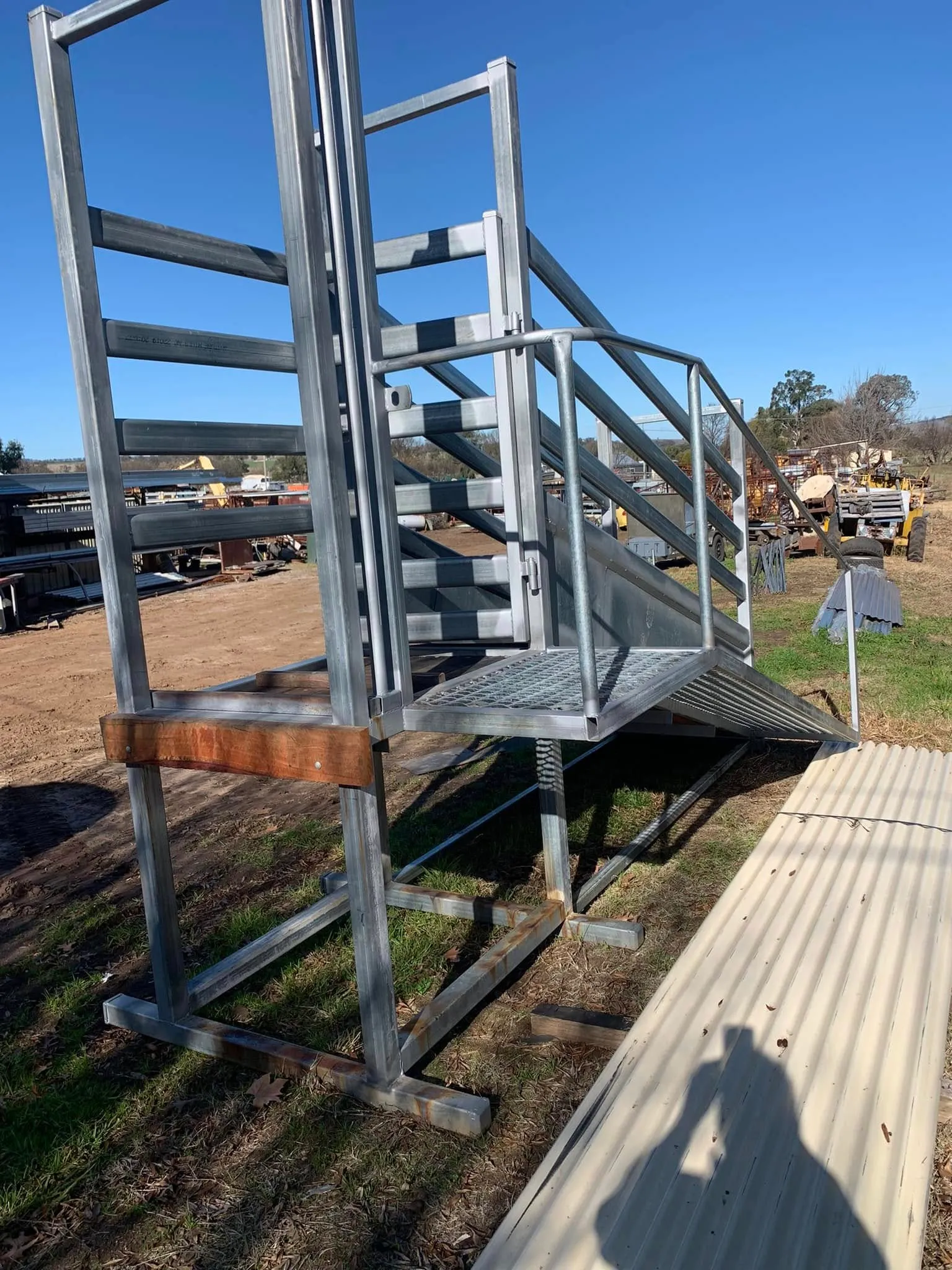 A galvanised livestock loading ramp with walkway, handrail and gate standing in the yard, with hills behind