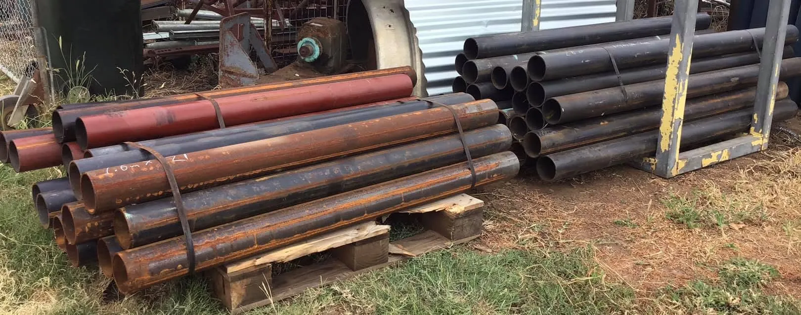 Stacks of large-diameter steel pipe on timber pallets in the yard, some red-primed, some black